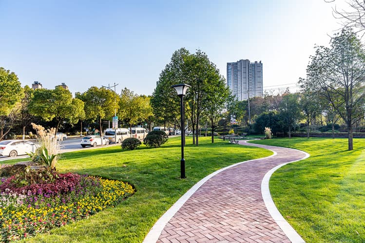 An image of a beautifully designed paver pathway cutting through a lush green city park with colorful flower beds lining the pathway.